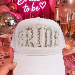 May include: A white trucker hat featuring the word BRIDE in large, silver sequined block letters across the front panel. The hat has a structured foam front, a mesh back, and a white cord trim detail running horizontally across the base of the crown.