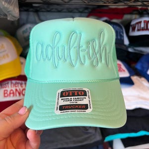 May include: A mint green trucker hat featuring the word adult-ish in raised script lettering on the front foam panel. A white sticker on the brim displays the text OTTO WORLD FAMOUS FOAM FRONT OSFM TRUCKER TERRY CLOTH SWEATBAND.