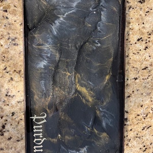 Puede incluir: Bandeja rectangular con un diseño veteado en negro, dorado y blanco. La palabra "Purdue" está escrita en una fuente decorativa en la esquina inferior izquierda. La bandeja tiene un acabado brillante.