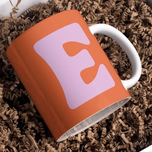 May include: A bright orange ceramic mug with a pink letter "E" printed on the front. The mug is sitting in brown packing peanuts.