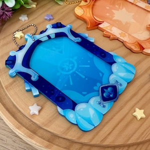 May include: Two decorative acrylic keychains. One is blue with a cloud and gem design, the other is orange with a star design. Both have a gold chain and are on a wooden surface with star-shaped embellishments.