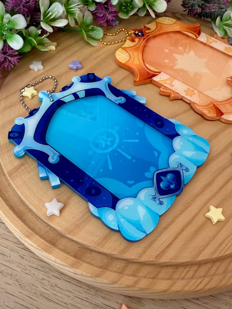 May include: Two decorative acrylic keychains. One is blue with a cloud and gem design, the other is orange with a star design. Both have a gold chain and are on a wooden surface with star-shaped embellishments.
