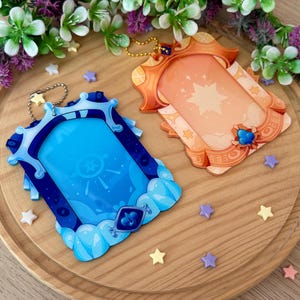 May include: Two decorative keychains with a fantasy theme. One is blue with a cloud design, the other is orange with a star design. Both have a rectangular frame and a gold-colored chain. Small star-shaped decorations are scattered around.