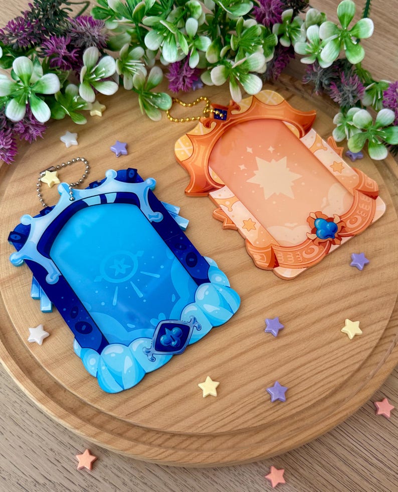 May include: Two decorative keychains with a fantasy theme. One is blue with a cloud design, the other is orange with a star design. Both have a rectangular frame and a gold-colored chain. Small star-shaped decorations are scattered around.