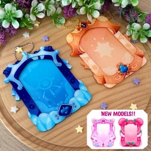 May include: Two decorative acrylic photo frames in blue and orange, with star and cloud designs. The blue frame has a silver chain, and the orange frame has a gold chain. The image also shows two pink frame designs with the text "NEW MODELS!!".