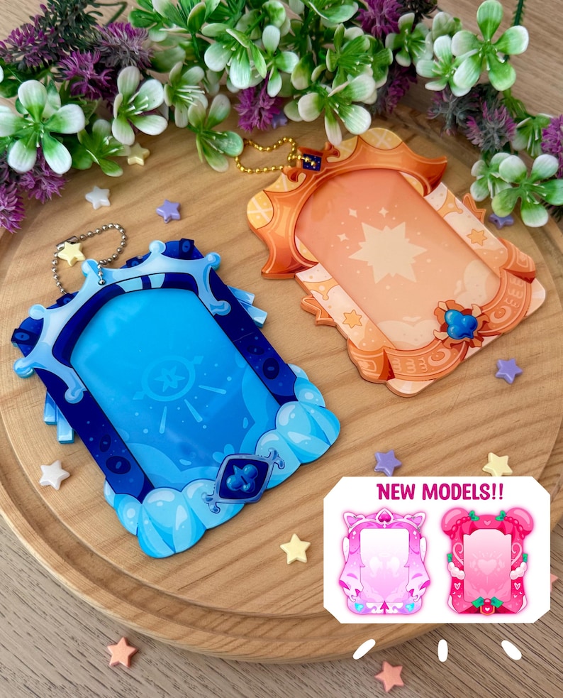 May include: Two decorative acrylic photo frames in blue and orange, with star and cloud designs. The blue frame has a silver chain, and the orange frame has a gold chain. The image also shows two pink frame designs with the text "NEW MODELS!!".