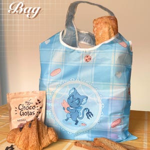 May include: A blue shopping bag with a plaid pattern, containing a loaf of bread. The bag has a cartoon illustration of a blue creature. A croissant, chocolate chips, and a bag of "Choco Gotas" are on a yellow tablecloth.