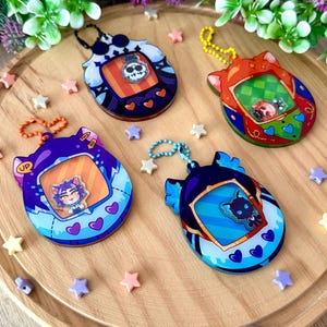 Può includere: Quattro portachiavi colorati a forma di Tamagotchi, ognuno con un personaggio diverso all'interno dello schermo. I portachiavi sono su una superficie di legno con piccole stelle sparse intorno.