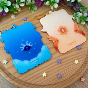 May include: Two acrylic keychains with gold chains. One is blue with a crown and eye design, the other is orange with a sun design. Both have the text "TYRENTOR OVOO". The keychains are on a wooden tray with star-shaped decorations.