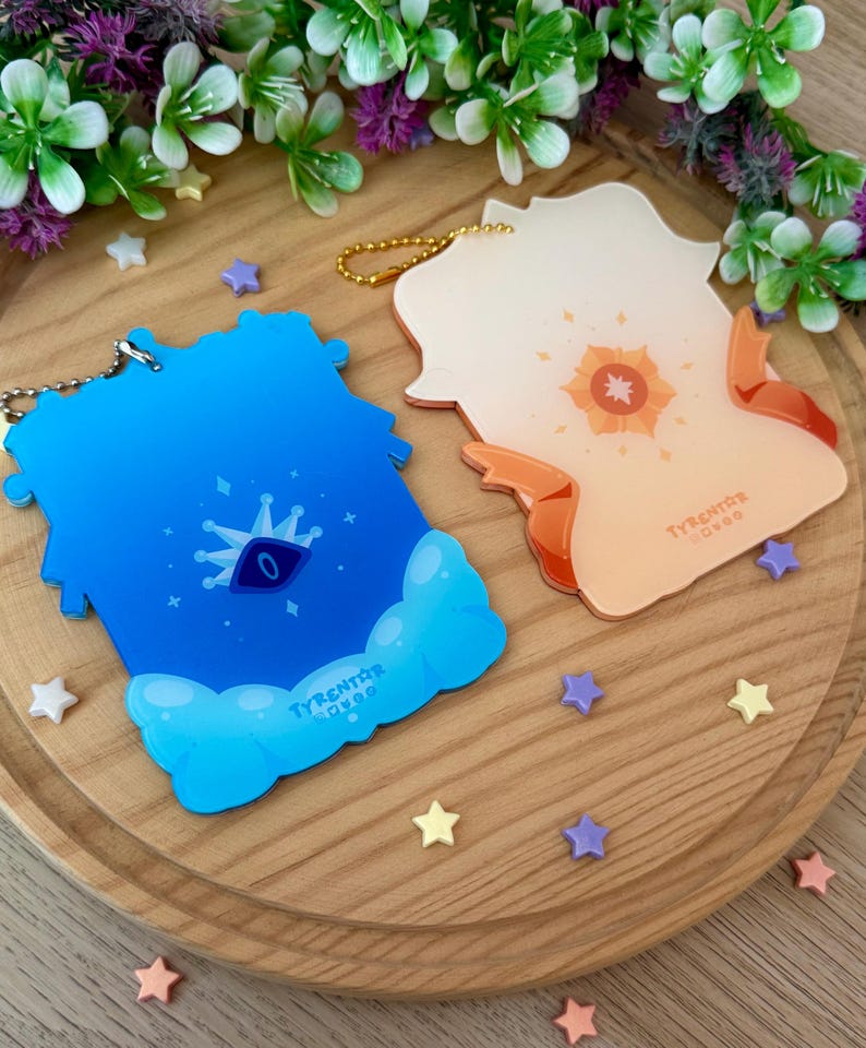 May include: Two acrylic keychains with gold chains. One is blue with a crown and eye design, the other is orange with a sun design. Both have the text "TYRENTOR OVOO". The keychains are on a wooden tray with star-shaped decorations.