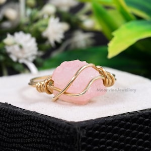 May include: A gold-toned wire-wrapped ring featuring a rough-cut, light pink gemstone. The gemstone is the focal point, secured by the wire that forms the ring's band. The ring is set against a soft, neutral background, highlighting the jewelry.