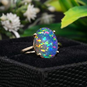 May include: An oval-shaped opal ring featuring a vibrant play of color with flashes of blue, green, and pink set in a gold band with four prongs. The text MoonvineJewellery is visible on the side of the ring band against a black velvet surface.