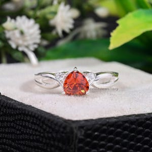 Può includere: Un anello d'argento con una pietra preziosa arancione a forma di pera come elemento centrale. Due pietre più piccole e chiare affiancano la pietra principale. La fascia dell'anello ha un design diviso, aggiungendo un tocco unico a questo gioiello.