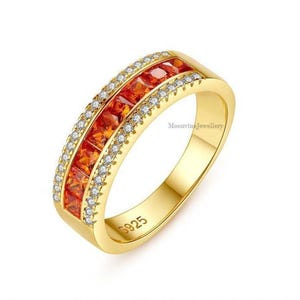 May include: A gold-toned ring featuring a central row of square-cut orange gemstones flanked by two rows of small, clear, round-cut stones. The inner band is engraved with the text S925 and the text MoonvineJewellery is visible on the side.