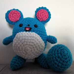 May include: A blue and white crocheted plush toy with pink ears and a white belly. The toy has a round body and a long tail.