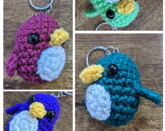 May include: Crocheted bird keychains in various colors: purple, green, blue, and teal. Each bird has a white belly, yellow beak, and black eyes. The keychains are on a wooden surface.