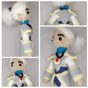 May include: A crocheted doll with white hair, blue eyes, and a blue and yellow outfit. The doll is wearing a necklace with a red gem pendant. The doll is handmade and has a detailed face.