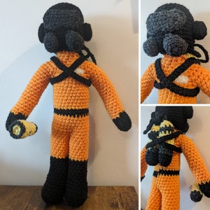 May include: A crocheted plush toy in the shape of a person wearing an orange jumpsuit with black accents and a black helmet. The toy is holding a yellow and orange flashlight in its right hand.