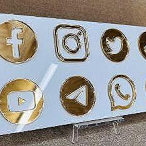 May include: A white rectangular sign displaying eight gold-coloured social media icons: Facebook, Instagram, Twitter, TikTok, YouTube, Telegram, WhatsApp, and LinkedIn. The icons are arranged in two rows of four.
