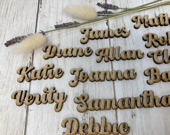 May include: Wooden name tags with names like James, Diane, Allan, Katie, Joanna, Verity, Samantha, and Debbie. The names are written in a cursive font and are arranged in a random pattern on a light wood surface.