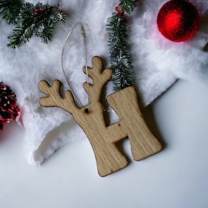 May include: Wooden reindeer ornament with the letter H cut out. The ornament is brown and has a string for hanging. It is sitting on a white fluffy surface.