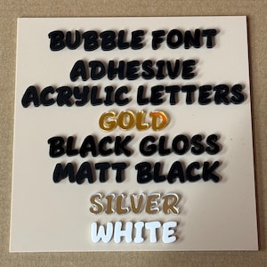 May include: A light tan acrylic board showcases adhesive acrylic letter samples. The text includes "Bubble Font," "Adhesive," "Acrylic Letters," "Gold," "Black Gloss," "Matt Black," "Silver," and "White." The letters are in black, gold, silver and white.