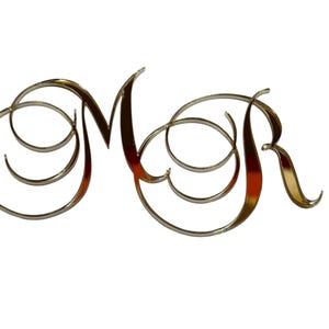 May include: A decorative metal monogram featuring the letters "M" and "R" in a flowing, cursive script. The letters are in a shiny, gold-coloured finish, with elegant swirls and curves. The background is plain white.