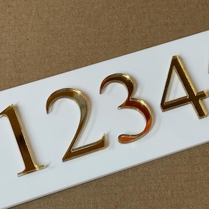 May include: Gold metallic numbers 1, 2, 3, 4, and 5 are affixed to a white rectangular surface. The numbers have a reflective, mirror-like finish, and are arranged in a horizontal row. The background is a light brown color.