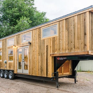 Puede incluir: Una casa pequeña de madera sobre ruedas con una ventana grande y puertas dobles. La casa está hecha de madera recuperada y tiene un aspecto rústico. La casa está sentada sobre un bastidor de remolque negro.