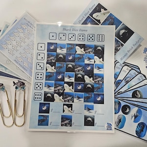 May include: A collection of shark-themed game components, including a game board, cards, and dice. The game board features a grid of shark images. Also included are decorative fish-shaped paper clips and a sticker with a dragon logo.
