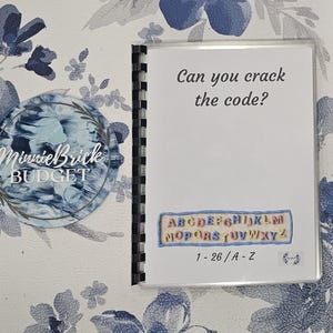 May include: A spiral-bound notebook with the text "Can you crack the code?" and an alphabet key. The notebook is on a floral patterned background with the text "MinnieBrick BUDGET" on a circular sticker.