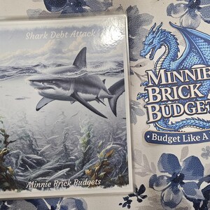 May include: A spiral-bound notebook with a cover featuring a shark illustration and the text "Shark Debt Attack." Next to it is a sticker with a blue dragon and the words "Minnie Brick Budgets" and "Budget Like A Dragon."