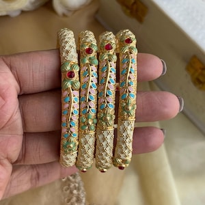 Puede incluir: Cuatro brazaletes dorados con intrincados diseños florales. Cada brazalete presenta una base crema con acentos rosas, turquesas y verdes. Los brazaletes están adornados con detalles de piedras preciosas rojas, creando un aspecto elegante y ornamentado. Los brazaletes se sostienen en una mano.