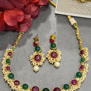 May include: A gold-toned necklace and matching stud earrings set featuring alternating round red and green cabochon stones, accented with small white seed pearls arranged in clusters along the metal chain and earring settings.