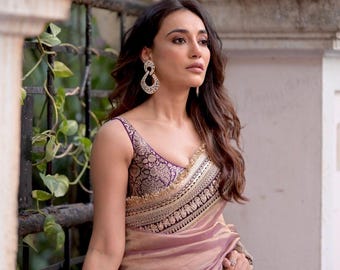 May include: A woman wearing a mauve saree with intricate gold embroidery and a matching purple blouse. The blouse has a deep V-neck and a detailed pattern. Large, ornate earrings complete the look. The saree is draped elegantly.