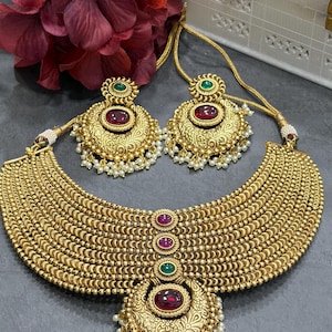 May include: A gold-toned necklace and matching earring set featuring intricate metalwork, embedded red and green cabochon stones, and small white pearl-like bead accents. The necklace consists of multiple rows of gold beads with a central pendant.