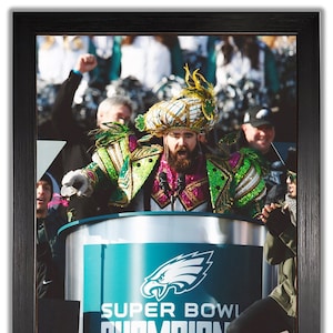 May include: Framed photograph of a person in a colorful costume speaking at a podium. The podium has the Philadelphia Eagles logo and the words "SUPER BOWL CHAMPIONS". The person is wearing a feathered headdress and a sequined jacket.