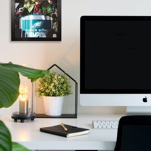 May include: A framed photograph of a Super Bowl celebration featuring the Philadelphia Eagles team. The image is displayed on a white wall above a desk with a computer, a notebook, and a decorative plant.