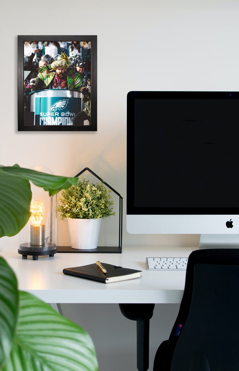 May include: A framed photograph of a Super Bowl celebration featuring the Philadelphia Eagles team. The image is displayed on a white wall above a desk with a computer, a notebook, and a decorative plant.