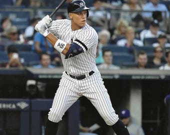 May include: A baseball player in a pinstriped uniform, wearing a black helmet with a white "NY" logo, stands at the plate holding a bat. The player is wearing black cleats and batting gloves. A baseball is visible in the foreground.