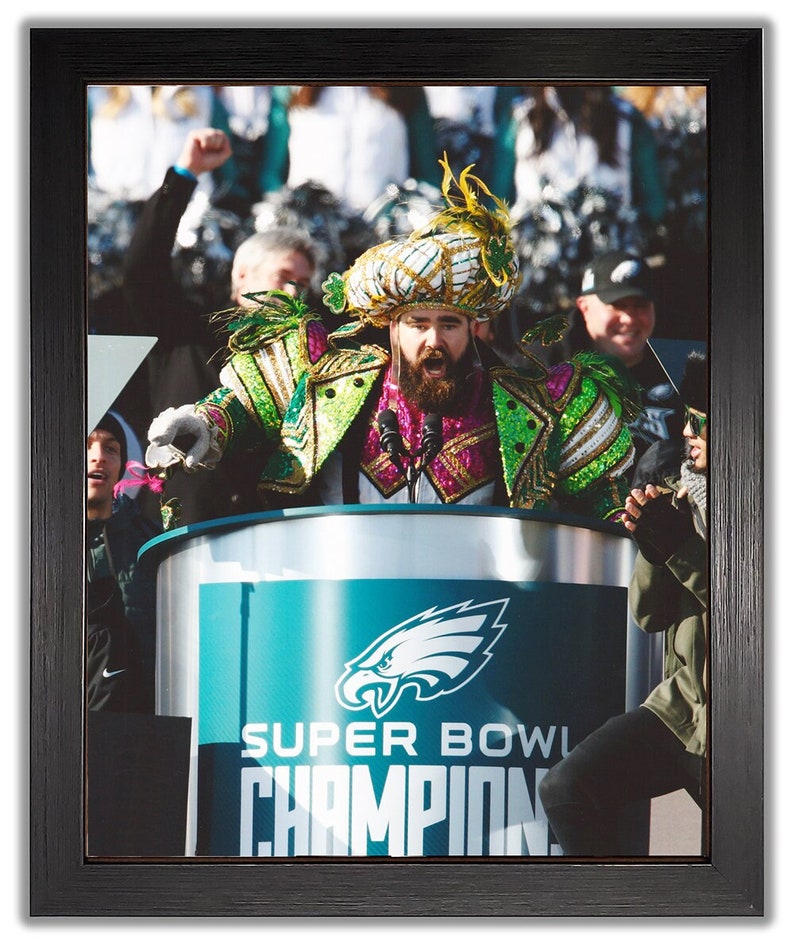 May include: Framed photograph of a person in a colorful costume speaking at a podium. The podium has the Philadelphia Eagles logo and the words "SUPER BOWL CHAMPIONS". The person is wearing a feathered headdress and a sequined jacket.