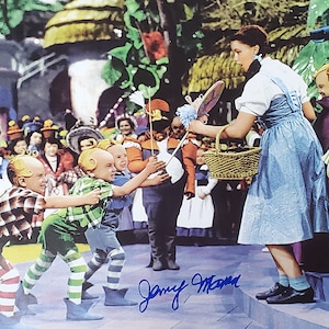 May include: A color image from the movie "The Wizard of Oz" featuring Judy Garland as Dorothy. The scene shows Dorothy with a basket, surrounded by Munchkins and other characters in a colorful, theatrical setting. The image includes an autograph.