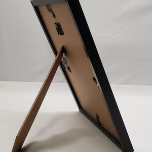 May include: A black picture frame with a brown backing and a wooden easel stand. The frame is rectangular and angled, showcasing its design. The frame is designed to display photos or artwork.