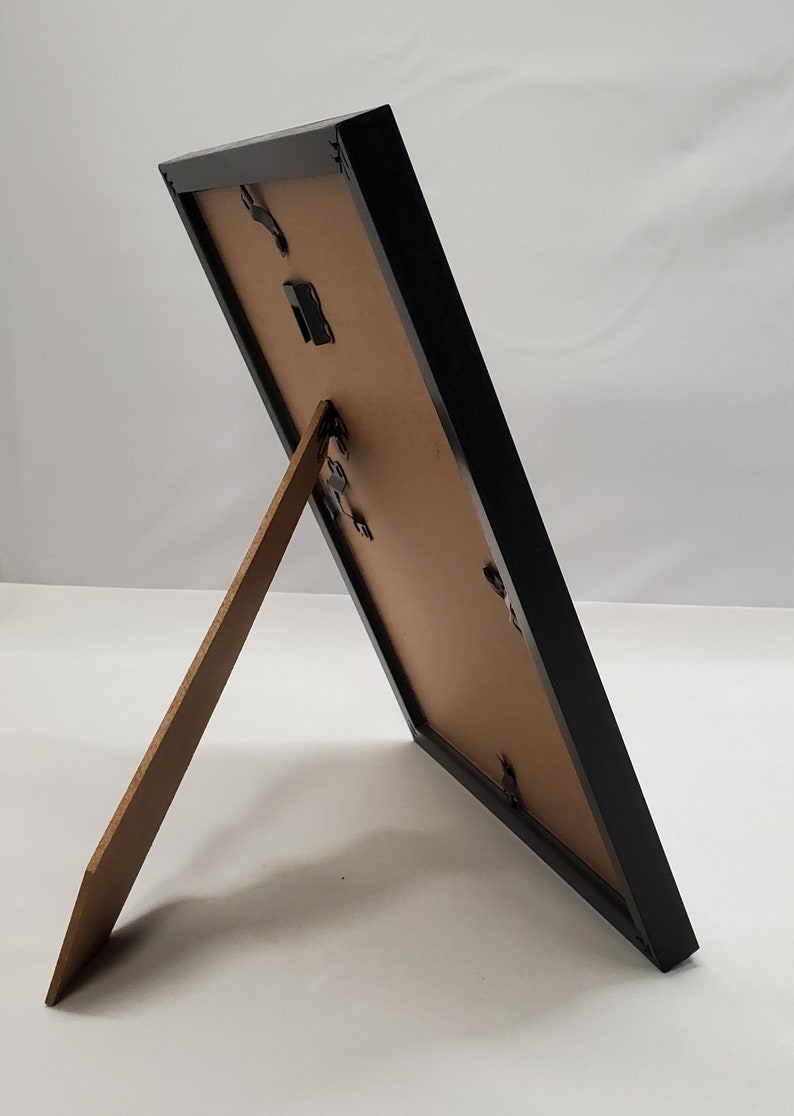 May include: A black picture frame with a brown backing and a wooden easel stand. The frame is rectangular and angled, showcasing its design. The frame is designed to display photos or artwork.