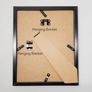 May include: Back of a black picture frame with a cardboard backing. The frame has metal hanging brackets and a cardboard stand. The text "Hanging Bracket" is printed on the cardboard.