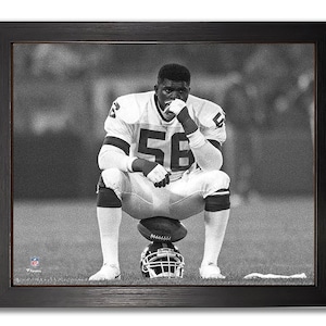 May include: Black and white framed photograph of a football player in a white and brown uniform with the number 56. The player is sitting on a football, with a helmet below. The player is resting his chin on his hand.