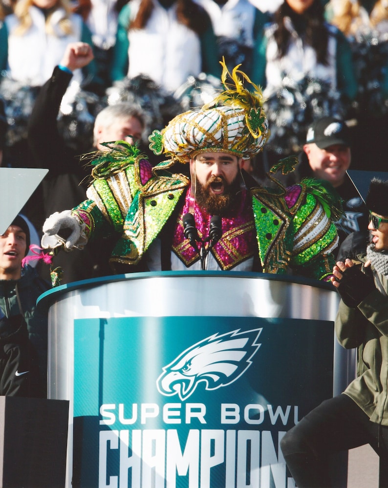 May include: A person in a colorful costume with a large headdress and a long beard speaks at a podium. The podium has the Philadelphia Eagles logo and the words "SUPER BOWL CHAMPIONS". The costume is green, pink, and gold.