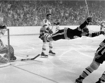Puede incluir: Fotografía en blanco y negro de un partido de hockey sobre hielo. Un jugador con uniforme negro y dorado celebra un gol, en el aire, con los brazos extendidos. Otro jugador está cerca. La multitud anima en el fondo. Un palo y un disco de hockey están sobre el hielo.
