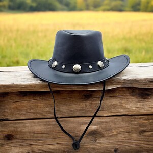 May include: A black leather cowboy hat with a silver band and decorative studs. The hat is hanging on a wooden fence.