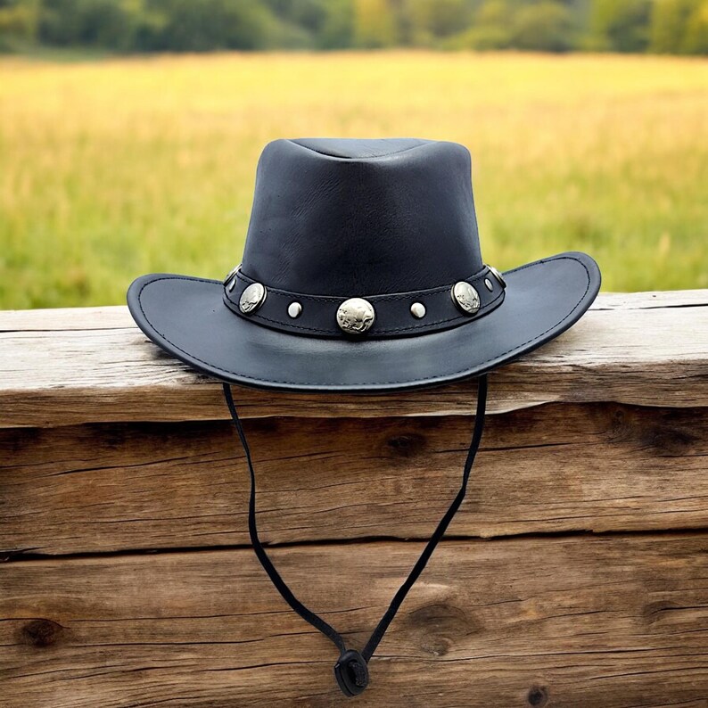 May include: A black leather cowboy hat with a silver band and decorative studs. The hat is hanging on a wooden fence.
